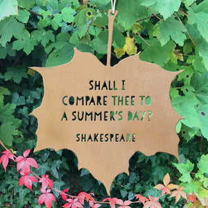 A leaf-shaped hanging sign with the quote 'SHALL I COMPARE THEE TO A SUMMER'S DAY? SHAKESPEARE' with lettering cut out of the metal, displayed outdoors against a background of green ivy and red autumn leaves.